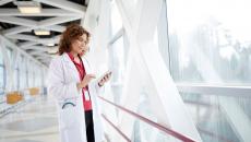 Healthcare provider holding a tablet while wearing a lab coat and standing in a hallway by a large window